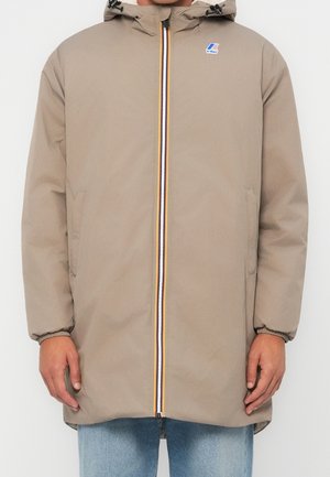 Beige hupparillinen takki, jossa edessä vetoketju ja sinisiä, valkoisia ja oransseja raitoja, joustavat hihansuut, sivutaskut, pidetty vaaleansinisten farkkujen päällä.