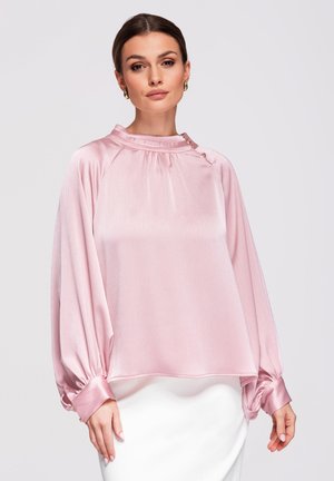 Femme aux cheveux foncés attachés en un chignon lisse, portant un chemisier ample et brillant de couleur rose pâle avec des manches longues et des boucles d'oreilles dorées, sur un fond clair.