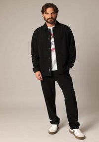 Zwarte button-up jas over een graphic T-shirt, gecombineerd met zwarte broeken en witte sneakers met grijze accenten. Gladde stof, nauwsluitende pasvorm.