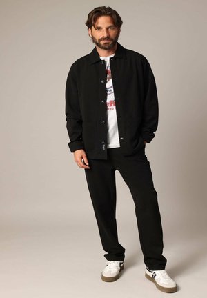 Veste noire à boutons sur un t-shirt graphique, associée à un pantalon noir et des baskets blanches avec des accents gris. Tissu lisse, coupe ajustée.