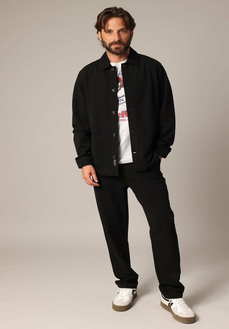 Zwarte button-up jas over een graphic T-shirt, gecombineerd met zwarte broeken en witte sneakers met grijze accenten. Gladde stof, nauwsluitende pasvorm.
