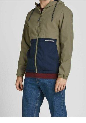 Man wearing olive green and navy blue zip-up hooded jacket with front pockets, blue jeans, and a maroon shirt underneath.