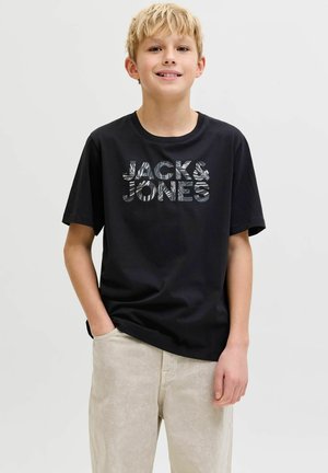 Blond boy standing with one hand in beige pants pocket, wearing a black T-shirt with patterned "JACK & JONES" text.
