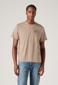 Beige színű pamut póló kerek nyakkal, rövid ujjakkal, a bal mellen egy kis zöld Levi's California logóval. Kék farmernadrággal viselve.
