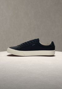 Baskets basses noires avec lacets noirs et semelle en caoutchouc blanche sur une surface claire avec un fond neutre uniforme.