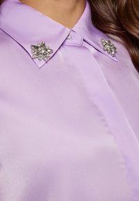 Lilla bluse med satinfinish, der har fremtrædende besætninger af juvelerede kraver formet som stjerner, som fremhæver udskæringen.