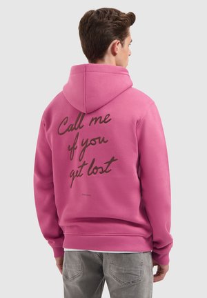 Man die met zijn rug naar de camera staat, gekleed in een roze hoodie met de kap opgezet en zwarte tekst op de rug die "Bel me als je bent verdwaald" zegt.