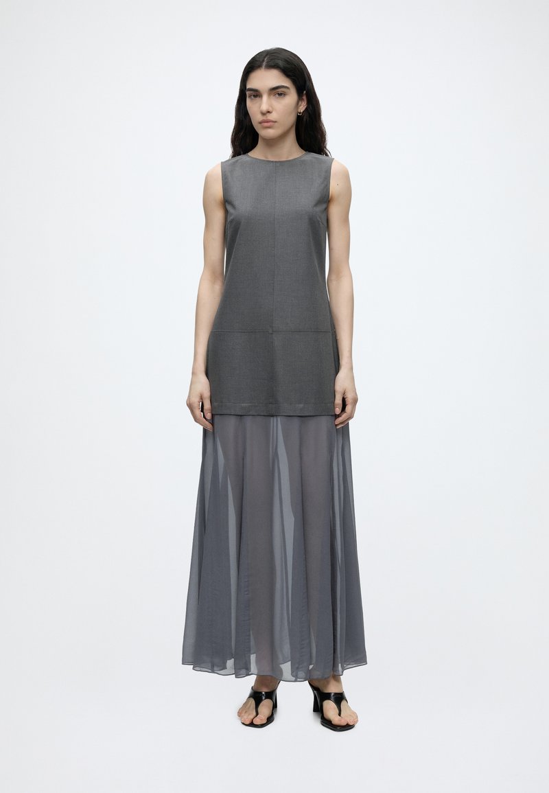 Femme aux longs cheveux foncés portant un haut gris sans manches et une jupe longue grise transparente, debout devant un fond clair uni.