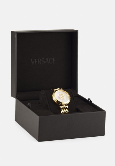 Zlaté hodinky Versace v čiernej krabičke s mäkkým interiérom. Majú okrúhly ciferník s textúrovaným povrchom a emblémom hlavy leva, s dizajnom náramku spojeného článku.