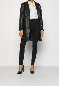 Abrigo de cuero negro de doble botonadura sobre una blusa de satén blanca, combinado con jeans ajustados negros y tacones negros puntiagudos.