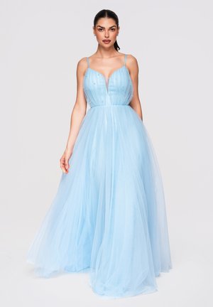 Femme en robe longue en tulle bleu clair avec fines bretelles et corsage orné, debout devant un fond clair uni.