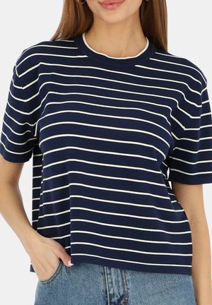 Kobieta ubrana w granatowo-biały paseczkowany, krótki rękaw z okrągłym dekoltem oraz jasnoniebieskie dżinsy.