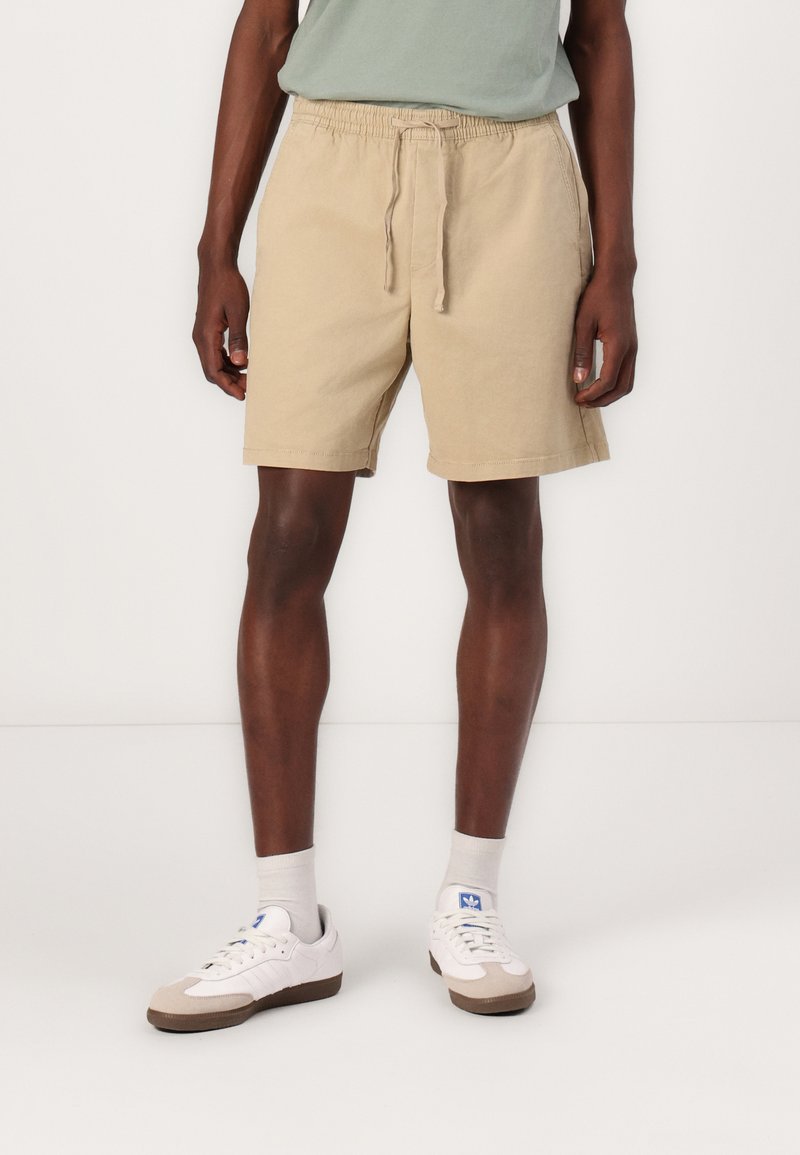 Beige bomulds shorts med elastisk talje, snor og sidelommer, parret med hvide sneakers og lysegrå sokker.