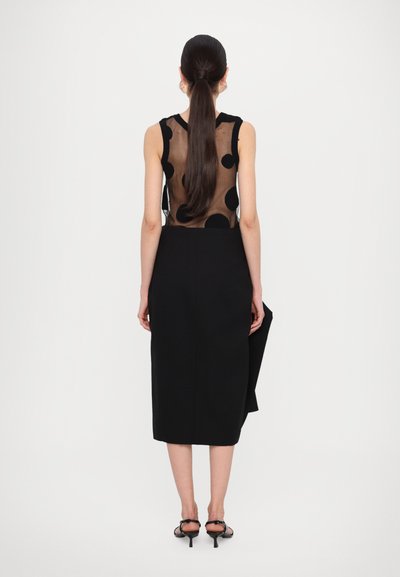 Black dress featuring a sheer back with polka dots, sleek silhouette, and a knee-length skirt. Model wears strappy black heels.