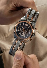Orologio in metallo argento e oro rosa con quadrante rotondo nero, tre sub-quadranti e cinturino grigio texturizzato, tenuto da una mano.