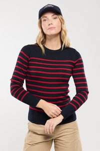Granatowy sweter w czerwone poziome paski, z prążkowaną strukturą, okrągłym dekoltem i guziczkami na ramionach. Noszony z beżowymi spodniami.