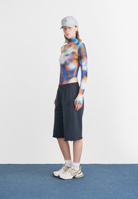 Pikkade käistega bodysuit, millel on värvikas gradient-muster, koos tumedate lühikestega. Aksessuaarid hõlmavad halli mütsi ja beeže tossusid.