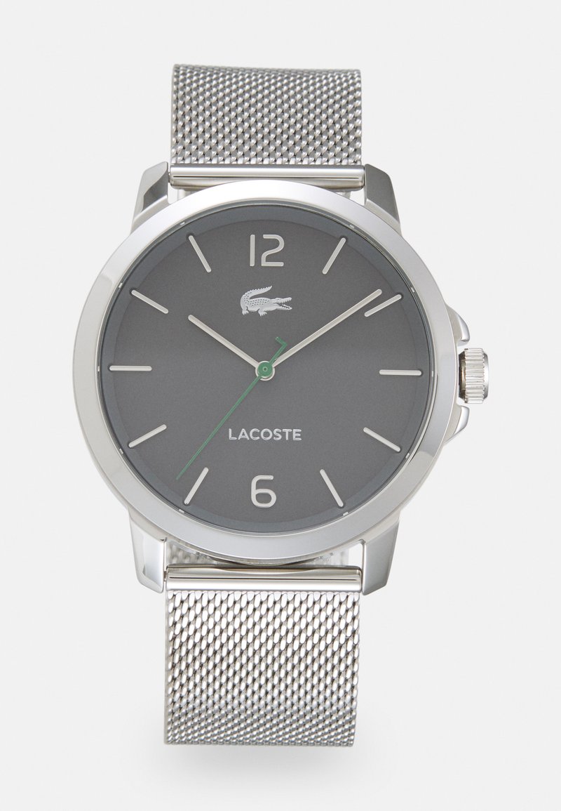 Reloj con dial gris y correa de malla plateada, que presenta una caja redonda de acero inoxidable, una manecilla de acento verde y marcadores de horas prominentes. Branding de Lacoste visible.