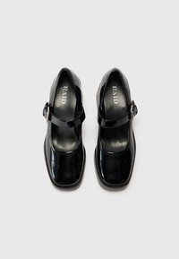 Black patent leather Mary Jane shoes with a square toe, adjustable ankle strap, and branded insole, featuring a smooth texture.