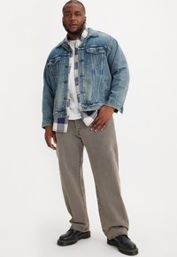 Spijkerjack met twee borstzakken en vervaagde blauwe kleur, gelaagd over een geruite overhemd en een wit t-shirt. Grijze wijde broek en zwarte laarzen.
