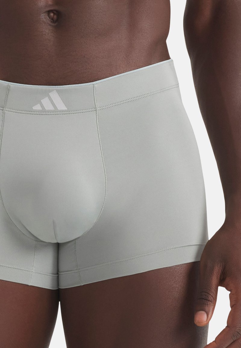 Torso masculin portant un boxer gris clair Adidas, avec le logo visible sur la ceinture et un tissu lisse avec des coutures apparentes.