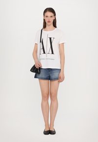 White cotton T-shirt with black "AX" logo, paired with denim shorts and black flats; model holds a small black crossbody bag.