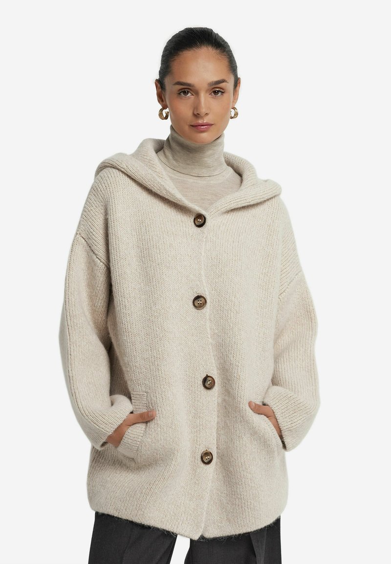 Beige stickad cardigan med huva, stora framfickor och fyra bruna knappar. Har en lös passform och textil struktur.