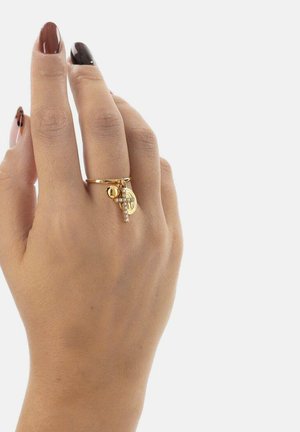 Main avec des ongles peints en rouge foncé brillant et noir, portant une bague en or ornée d'une croix, d'une pièce de monnaie et d'un charm rond sur le majeur, sur fond blanc.