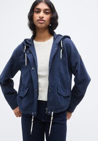 Μπλε σκούρο ελαφρύ μπουφάν με κουκούλα, κλείσιμο με κορδόνι, εμπρόσθιες τσέπες και αντίθετα κορδόνια. Φορεμένο πάνω από ένα υφασμάτινο λευκό τοπ.