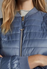 Personne portant une veste matelassée bleu clair avec une fermeture éclair devant, un col et des manches gris clair, avec de longs cheveux blonds visibles.