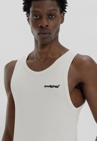 White ribbed tank top with a crew neck, featuring a small black logo on the left side, fitted design, and wide shoulder straps.