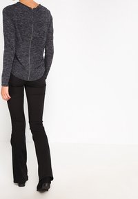 Suéter de punto gris con cierre de cremallera en la parte posterior, combinado con pantalones de campana negros. La tela parece tener textura, creando un aspecto en capas.