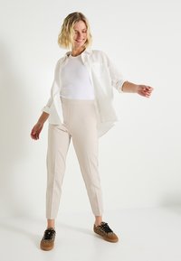 Chemise blanche à boutons superposée sur un débardeur blanc ajusté, associée à un pantalon beige tailleur et des baskets marron à motif léopard.