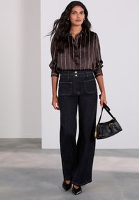 Chemise boutonnée en satin à rayures marron associée à un pantalon en denim foncé taille haute avec poches avant, souligné par des boutons dorés.