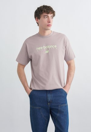 Noormees lokkis juustega, kes kannab helelillat New Balance Athletics t-särki ja siniseid teksaseid, seisab käed taskutes ja vaatab vasakule.