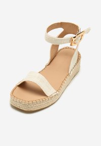 Beige espadrillesandal med et vævet stofoverdel, ankelrem med en guldbeslået spænde og en mellemsål indpakket i jute.
