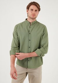 Chemise verte à manches longues avec col montant, fermeture à boutons et poignets retroussés. Fabriquée en tissu léger avec une texture subtile.