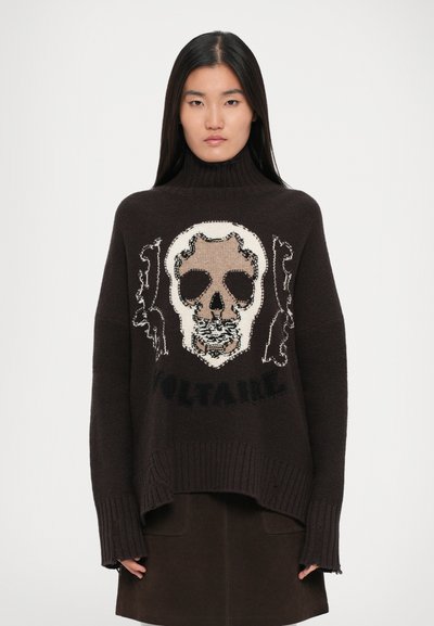 Mujer joven con cabello largo y negro, vistiendo un suéter de cuello alto oscuro que presenta una gran calavera y el texto "VOLTAIRE", combinado con una falda oscura.