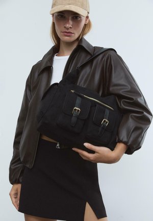 Jeune femme portant une casquette beige, une veste en cuir marron foncé, une jupe noire avec une fente, tenant un grand sac à main noir avec des poches frontales et des boucles.