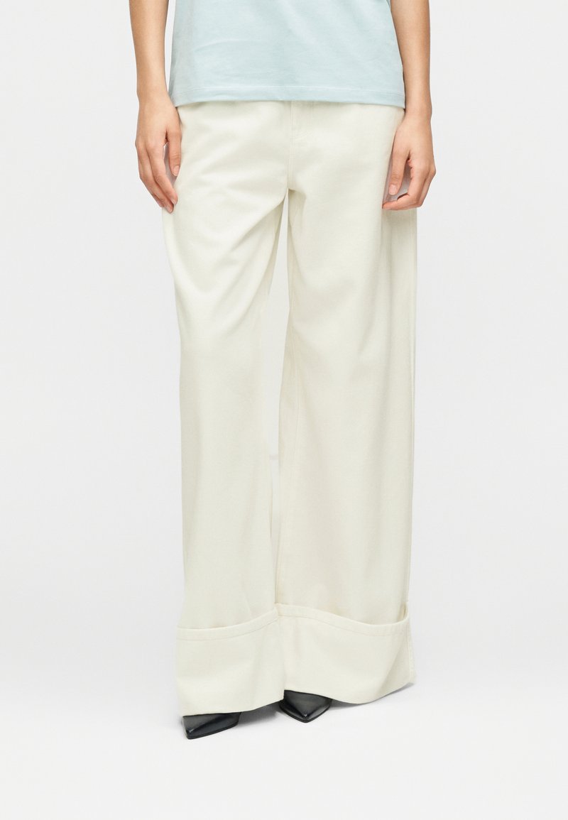 Παντελόνι με φαρδιά μπατζάκια από απαλό, εκρού ύφασμα με λεία υφή, διακοσμημένο με διπλωμένο τελείωμα και συνδυασμένο με μαύρες μυτερές γόβες.