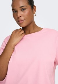 Lyserød bomuldstop med afslappet pasform, bred halsudskæring og korte ærmer. Modellen har en tatovering synlig på håndleddet og bærer øreringe i hoop-stil.