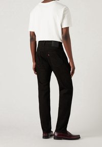 Personne debout, portant un jean noir, une chemise blanche à manches courtes et des mocassins en cuir marron foncé, vue de dos sur un fond uni.
