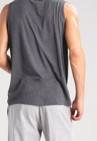 Muž v černém bezrukávovém tričku a světle šedých šortkách, stojící zády k fotoaparátu na jednobarevném pozadí.