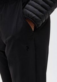 Χέρι στην τσέπη μαύρου παντελονιού, με ορατό μανίκι μαύρου πλεκτού μπουφάν.