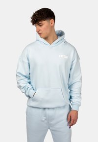 Hellblauem Hoodie in lockerer Passform, mit einer Fronttasche und weißem Logo. Kombiniert mit passenden hellblauen Jogginghosen, die einen kleinen farblichen Akzent aufweisen.
