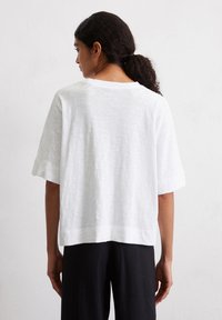 Un haut blanc à manches courtes présente une coupe décontractée, un tissu texturé, une encolure large et des manches roulées, associé à un pantalon noir.