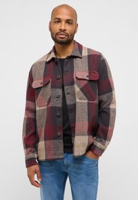 Karierte Flanellhemd in Rot, Beige und Dunkelgrau mit zwei Brusttaschen, Knopfverschluss und lockerem Sitz, kombiniert mit einem schwarzen Shirt und blauer Jeans.