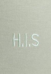 Helles grünes Gewebe mit gestickten Buchstaben "H.I.S" in einem etwas dunkleren Grün, mit einer glatten, strukturierten Oberfläche.
