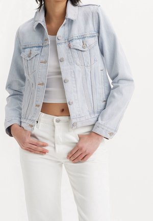 Femme portant une veste en jean bleu clair sur un crop top blanc et un pantalon blanc taille haute, mains posées sur les hanches, fond uni.