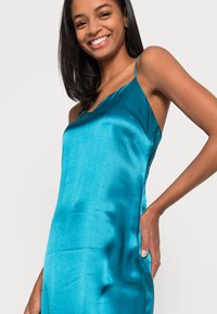 Vestido slip em seda teal com alças finas, decote em V e um corte descontraído. Textura suave e detalhes de bolsos laterais.